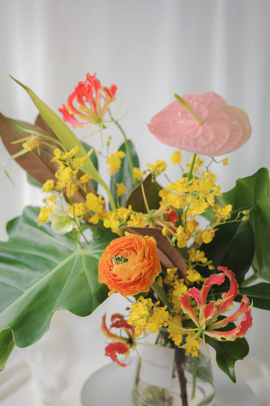 Tropical-inspired seasonal flower arrangement with vibrant blooms like ranunculus, anthuriums, gloriosa lilies, and lush foliage in a glass vase.