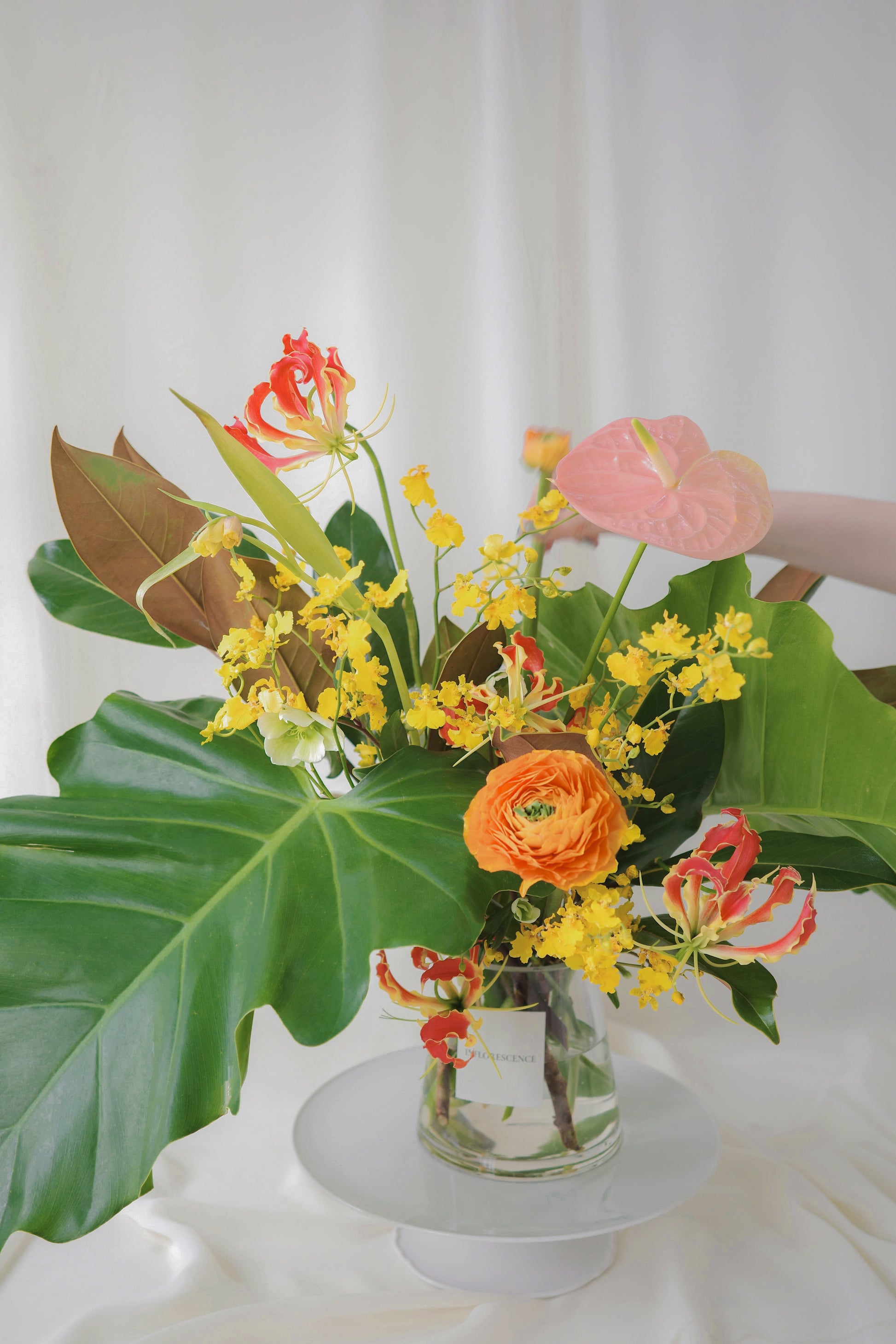 Tropical-inspired seasonal flower arrangement with vibrant blooms like ranunculus, anthuriums, gloriosa lilies, and lush foliage in a glass vase.