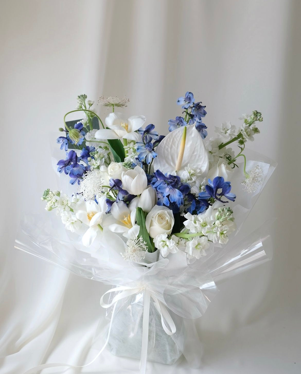 Elegant white and blue bouquet with delphiniums, tulips, and roses by Studio Inflorescence Singapore